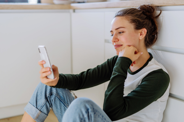 Young woman sitting with smartphone in her kitchen.