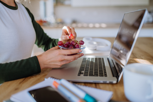 Side view of woman holding bowl with grapes above a desk with computer, diary and smartphone. Work-life balance concept.