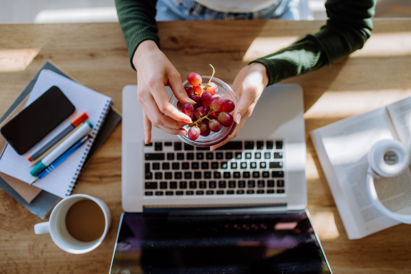 Top view of woman holding bowl with grapes above a desk with computer, diary and smartphone. Work-life balance concept.