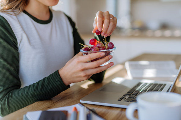 Side view of woman holding bowl with grapes above a desk with computer, diary and smartphone. Work-life balance concept.