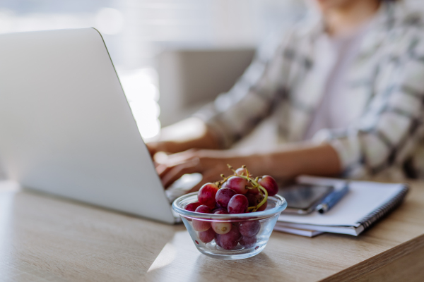 Side view of woman holding bowl with grapes above a desk with computer, diary and smartphone. Work-life balance concept.