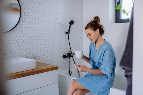 Young woman in bathroom with phone and cup of coffee, morning routine and relaxation concept.