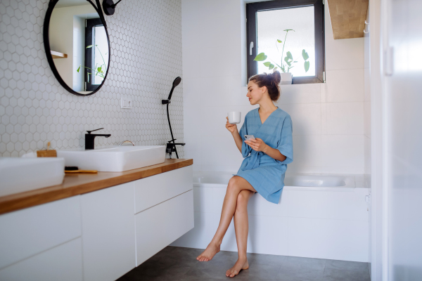 Young woman in bathroom with phone and cup of coffee, morning routine and relaxation concept.