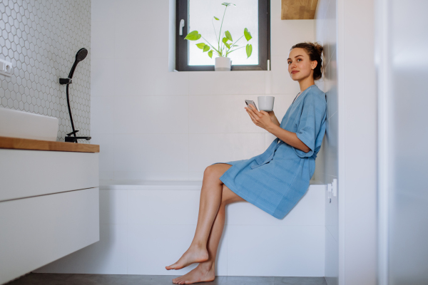 Young woman in bathroom with phone and cup of coffee, morning routine and relaxation concept.