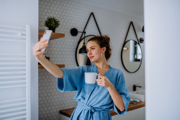 Young woman taking selfie in bathroom, enjoying a cup of coffee.