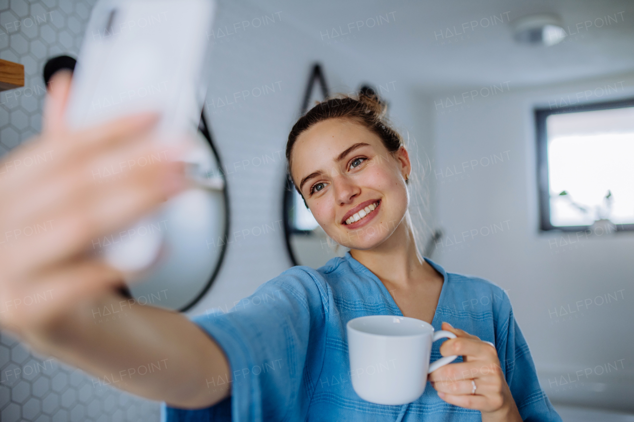 Young woman taking selfie in bathroom, enjoying a cup of coffee.