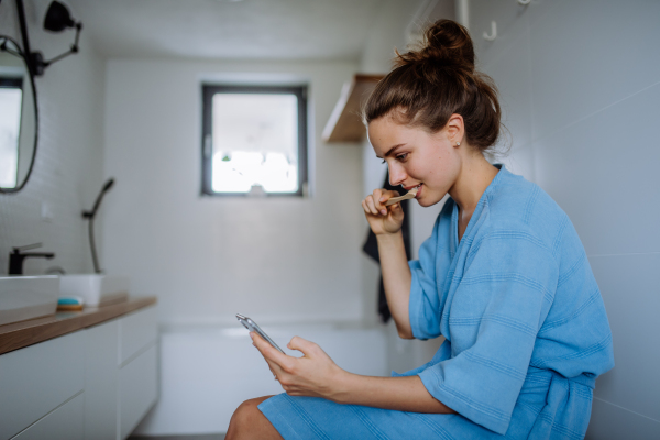 Young woman brushing teeth and scrolling her smartphone, morning routine concept.