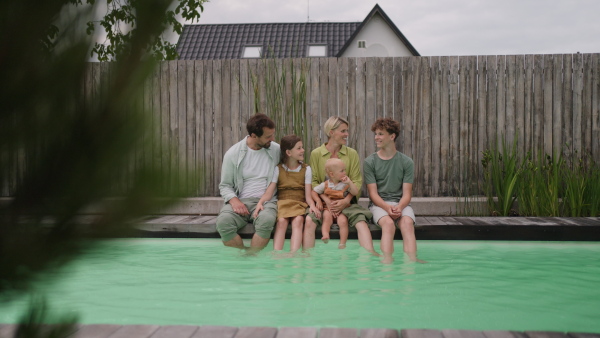 A video of beautiful young family with three kids sitting in pool with legs in water.