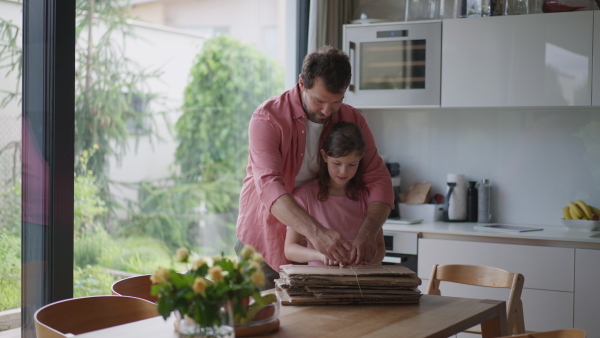 A father and girl preparing cardboard boxes for recycling, working in kitchen together. Sustainable living.