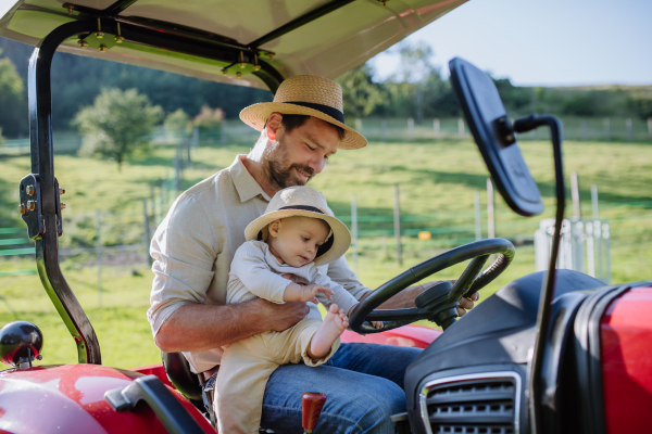 Farmer father riding tractor with his little baby son. Little baby growing up on family farm. Concept of multigenerational farming.