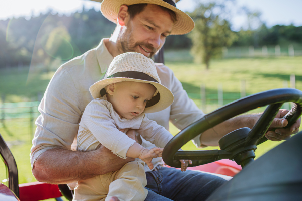 Farmer father riding tractor with his little baby son. Little baby growing up on family farm. Concept of multigenerational farming.