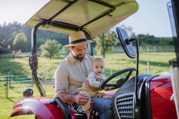 Farmer father riding tractor with his little baby son. Little baby growing up on family farm. Concept of multigenerational farming.