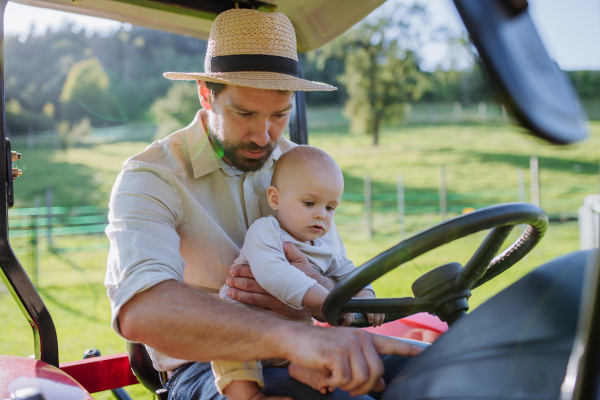 Farmer father riding tractor with his little baby son. Little baby growing up on family farm. Concept of multigenerational farming.