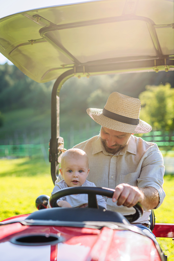 Farmer father riding tractor with his little baby son. Little baby growing up on family farm. Concept of multigenerational farming.