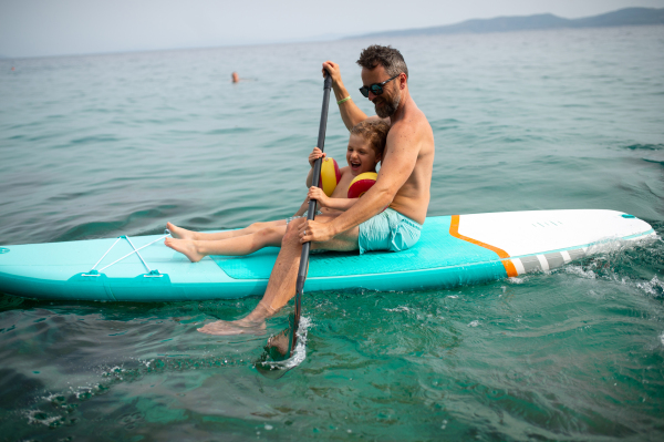 A happy father and little son on paddle board in sea together. Fatherhood and quality time with child concept.