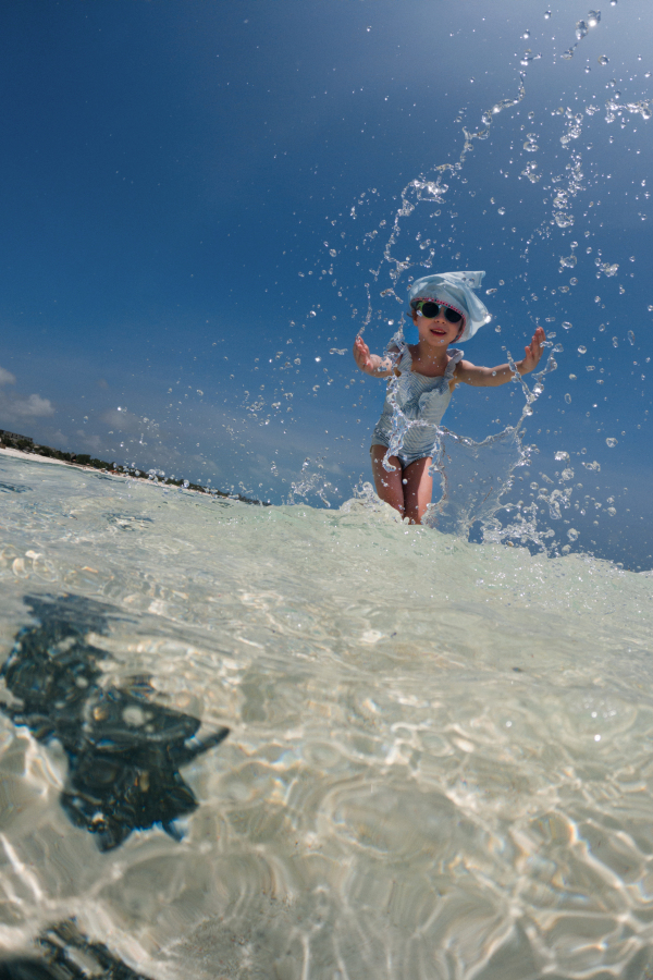 Portrait of a little girl splashing sea water on the beach. Low angle, full body shot of smilling girl in blue swimsuit, enjoying sandy beach and crystalline sea of Mnemba beach in Zanzibar, with copy space.. Concept of beach summer vacation.Concept of beach summer vacation with kids.