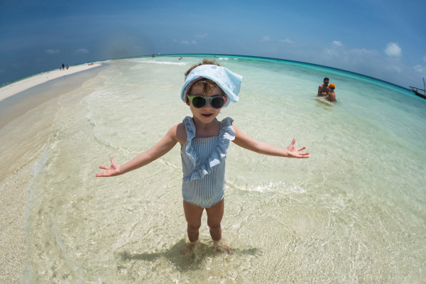 Portrait of a little girl standing seawater on the beach. Fisheye, full body shot of smilling girl in blue swimsuit, enjoying sandy beach and crystalline sea of Mnemba beach in Zanzibar, with copy space.. Concept of beach summer vacation.Concept of beach summer vacation with kids.