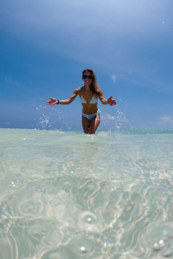 Portrait of a beautiful slim woman standing in sea in bikini and splashing water. Low angle, full body shot of woman in blue swimsuit, sunglasses enjoying sandy beach and crystalline sea of Mnemba beach in Zanzibar. Concept of beach summer vacation.