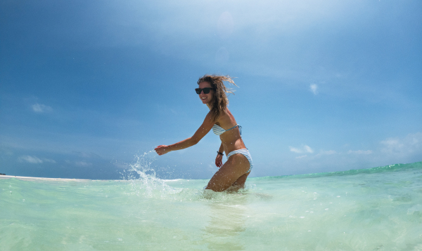 Portrait of a beautiful slim woman standing in sea in bikini and splashing water. Low angle, full body shot of woman in blue swimsuit, sunglasses enjoying sandy beach and crystalline sea of Mnemba beach in Zanzibar. Concept of beach summer vacation.