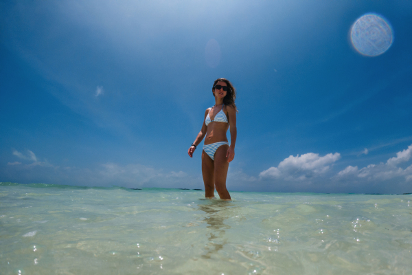 Portrait of a beautiful slim woman standing in sea water on beach in bikini. Low angle, full body shot of woman in blue swimsuit, sunglasses enjoying sandy beach and crystalline sea of Mnemba beach in Zanzibar, with copy space. Concept of beach summer vacation.