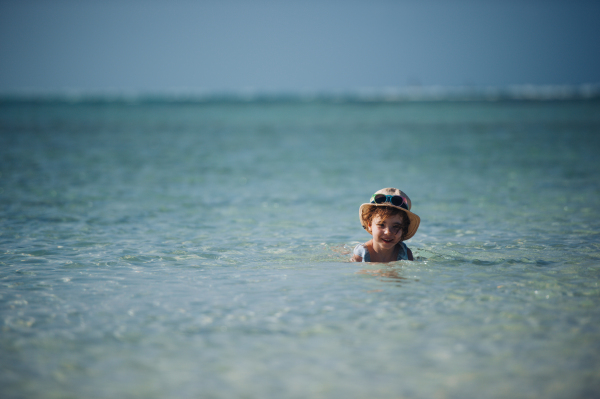 Portrait of a little girl swimming in a sea with straw hat on head. Full body shot of smilling girl enjoying turquoise seawater on Mnemba beach in Zanzibar, with copy space. Concept of beach summer vacation with kids.