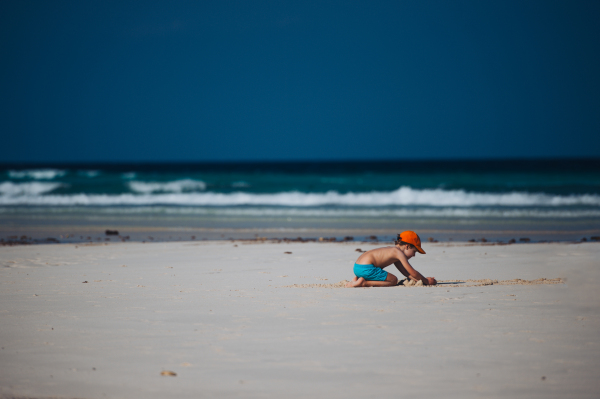 Portrait of a little boy playing on the beach, building sand castle. Full body shot of young boy in blue swimsuit kneeling, enjoying sandy beach and crystalline sea of Mnemba beach in Zanzibar, with copy space. Concept of beach summer vacation with kids.