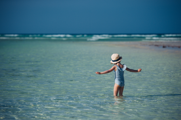 Rear view of a beautiful little girl standing in the water on the beach in a swimsuit and a straw hat. Full body shot of smilling girl in blue swimsuit, enjoying sandy beach and crystalline sea of Mnemba beach in Zanzibar, with copy space. Concept of beach summer vacation with kids.