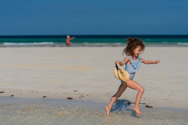 Side view of a little girl running on the beach in a swimsuit and a straw hat in hand. Full body shot of smilling girl in blue swimsuit, enjoying sandy beach and crystalline sea of Mnemba beach in Zanzibar, with copy space. Concept of beach summer vacation with kids.