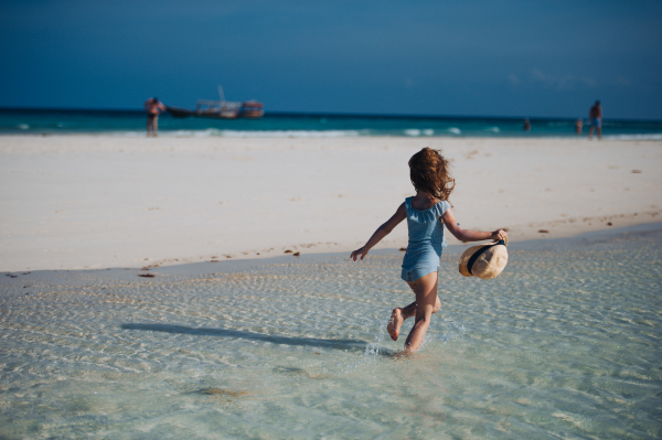 Rear view of a little girl running on the beach in a swimsuit and a straw hat in hand. Full body shot of smilling girl in blue swimsuit, enjoying sandy beach and crystalline sea of Mnemba beach in Zanzibar, with copy space. Concept of beach summer vacation with kids.