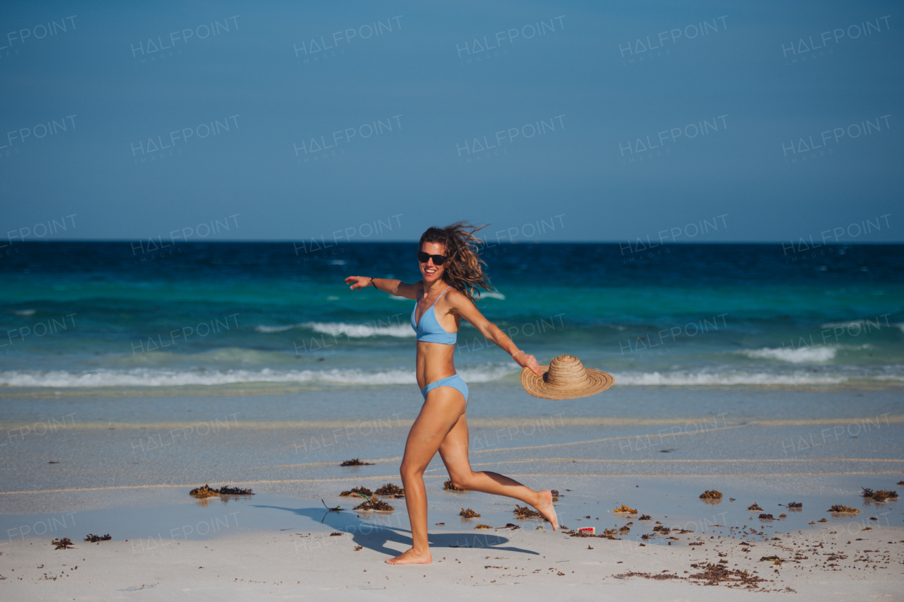 Portrait of a beautiful slim woman running on the beach in a bikini and a straw hat, enjoying sunbeams. Full body shot of woman in blue swimsuit, sunglasses enjoying sandy beach and crystalline sea of Mnemba beach in Zanzibar. Concept of beach summer vacation.