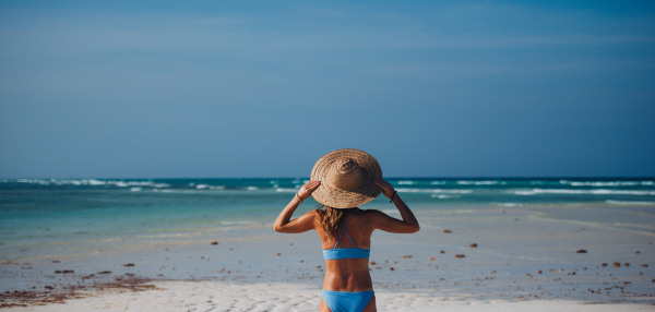 Rear view of a beautiful slim woman standing on the beach in a bikini and a straw hat. Waist up shot of woman in blue swimsuit enjoying sandy beach and crystalline sea of Mnemba beach in Zanzibar. Concept of beach summer vacation. Banner with copyspace.