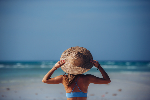 Rear view of a beautiful slim woman standing on the beach in a bikini and a straw hat. Waist up shot of woman in blue swimsuit enjoying sandy beach and crystalline sea of Mnemba beach in Zanzibar, with copy space. Concept of beach summer vacation.