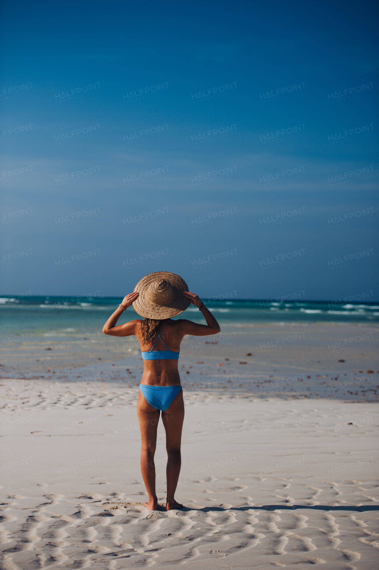 Rear view of a beautiful slim woman standing on the beach in a bikini and a straw hat. Full body shot of woman in blue swimsuit enjoying sandy beach and crystalline sea of Mnemba beach in Zanzibar. Concept of beach summer vacation.