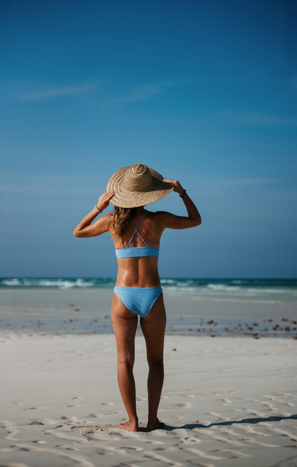 Rear view of a beautiful slim woman standing on the beach in a bikini and a straw hat. Full body shot of woman in blue swimsuit enjoying sandy beach and crystalline sea of Mnemba beach in Zanzibar. Concept of beach summer vacation.