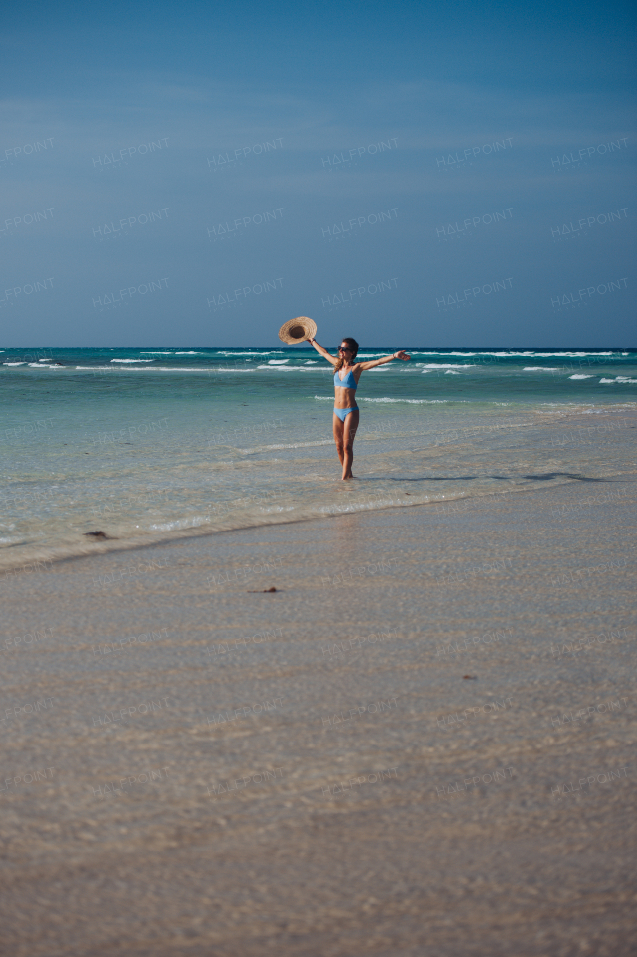 Portrait of a beautiful slim woman standing on the beach in a bikini and a straw hat, enjoying sunbeams. Full body shot of woman in blue swimsuit, sunglasses enjoying sandy beach and crystalline sea of Mnemba beach in Zanzibar. Concept of beach summer vacation.