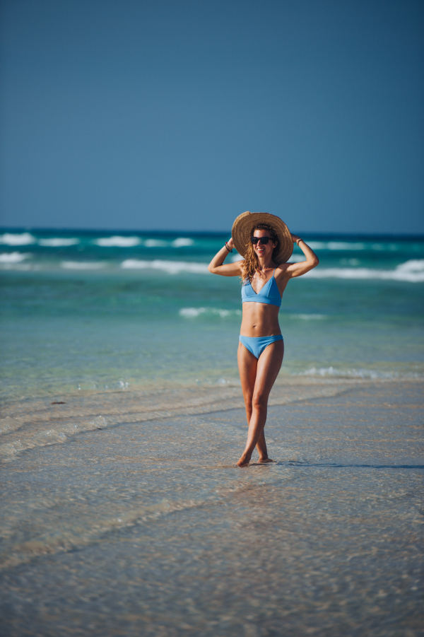 Portrait of a beautiful slim woman standing on the beach in a bikini and a straw hat. Full body shot of woman in blue swimsuit, sunglasses enjoying sandy beach and crystalline sea of Mnemba beach in Zanzibar. Concept of beach summer vacation.