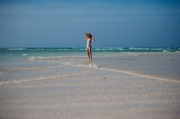 Portrait of a beautiful slim woman standing on the beach in a bikini and a straw hat, enjoying sunbeams. Full body shot of woman in blue swimsuit, sunglasses enjoying sandy beach and crystalline sea of Mnemba beach in Zanzibar. Concept of beach summer vacation.