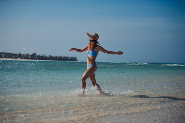 Portrait of a beautiful slim woman splashing water on the beach in a bikini and a straw hat. Full body shot of woman in blue swimsuit, sunglasses enjoying sandy beach and crystalline sea of Mnemba beach in Zanzibar. Concept of beach summer vacation.