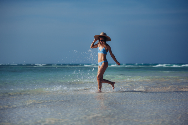 Portrait of a beautiful slim woman splashing water on the beach in a bikini and a straw hat. Full body shot of woman in blue swimsuit, sunglasses enjoying sandy beach and crystalline sea of Mnemba beach in Zanzibar. Concept of beach summer vacation.