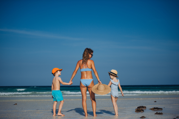 Rear view of a mother with little girl and boy walking on the beach. Full body shot of family in swimsuit, enjoying sandy beach and crystalline sea of Mnemba beach in Zanzibar, with copy space. Concept of beach summer vacation with kids.