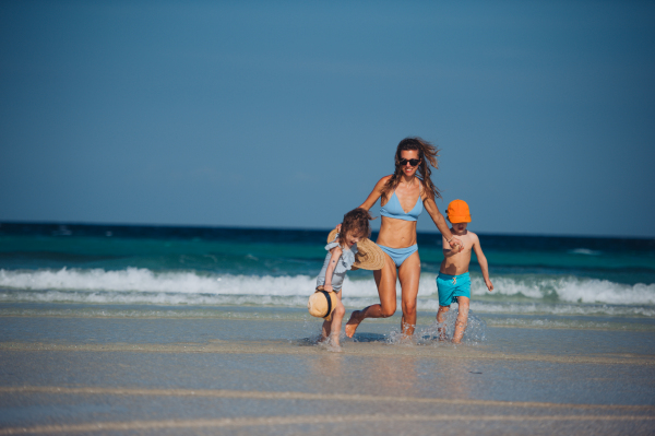 Young mother with little girl and boy walking on the beach. Full body shot of family in swimsuit, enjoying sandy beach and crystalline sea of Mnemba beach in Zanzibar, with copy space. Concept of beach summer vacation with kids.