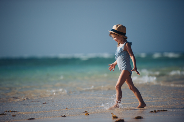 Portrait of a little girl walking on the beach in a swimsuit and a straw hat. Full body shot of smilling girl in blue swimsuit, enjoying sandy beach and crystalline sea of Mnemba beach in Zanzibar, with copy space. Concept of beach summer vacation with kids.