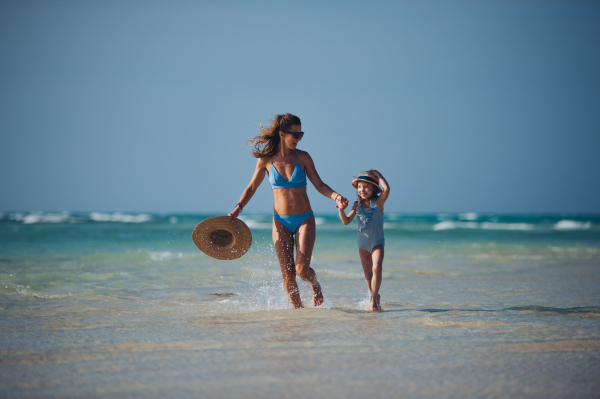 Portrait of a mother with little daughter walking on the beach. Full body shot of family in swimsuit, enjoying sandy beach and crystalline sea of Mnemba beach in Zanzibar, with copy space. Concept of beach summer vacation with kids.