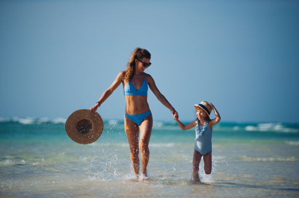 Portrait of a mother with little girl walking on the beach. Full body shot of family in swimsuit, enjoying sandy beach and crystalline sea of Mnemba beach in Zanzibar, with copy space. Concept of beach summer vacation with kids.