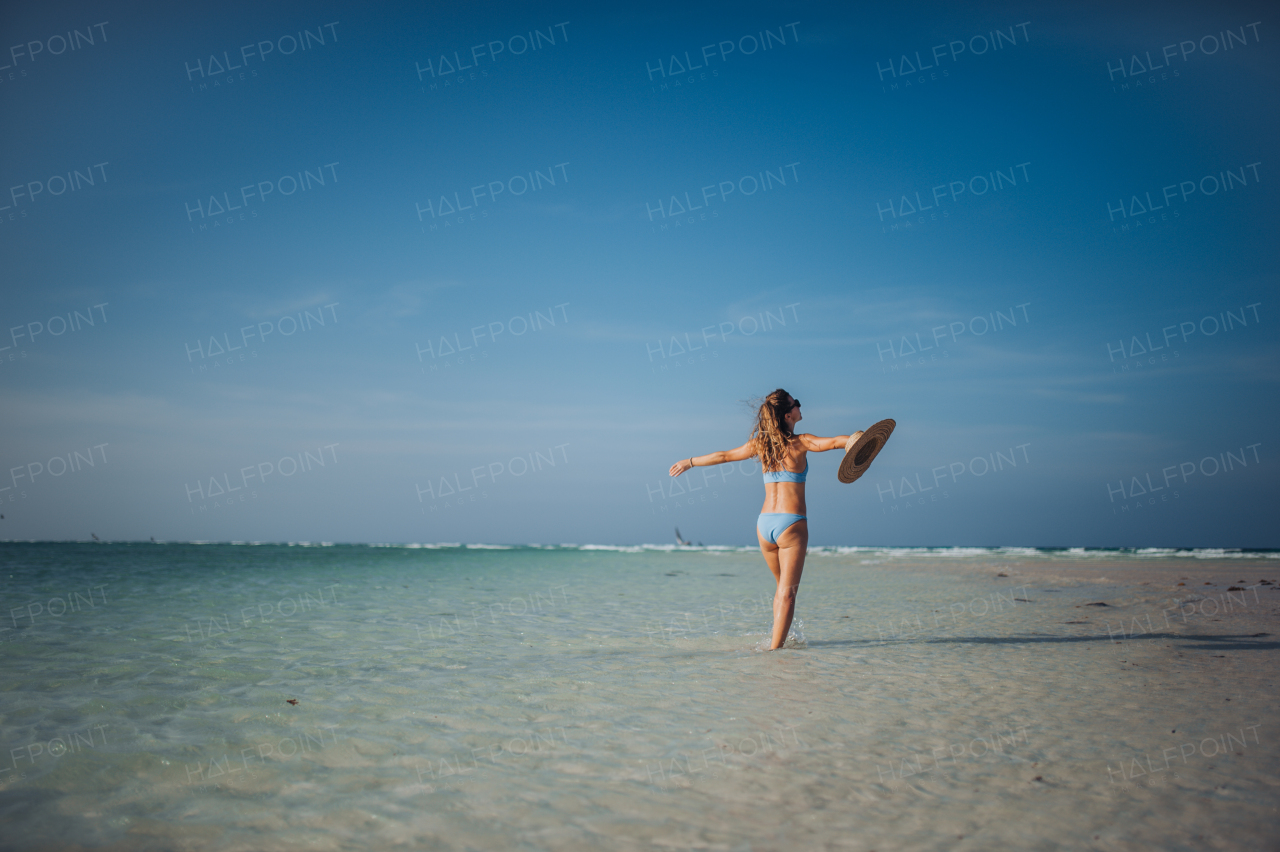 Rear view of a beautiful slim woman standing on the beach in a bikini and a straw hat, enjoying sunbeams. Full body shot of woman in blue swimsuit, sunglasses enjoying sandy beach and crystalline sea of Mnemba beach in Zanzibar. Concept of beach summer vacation.