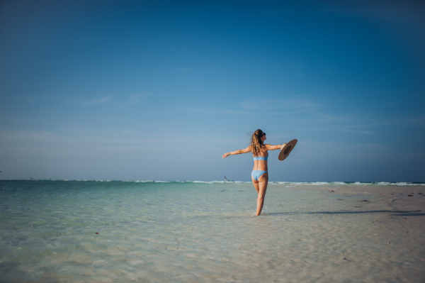 Rear view of a beautiful slim woman standing on the beach in a bikini and a straw hat, enjoying sunbeams. Full body shot of woman in blue swimsuit, sunglasses enjoying sandy beach and crystalline sea of Mnemba beach in Zanzibar. Concept of beach summer vacation.