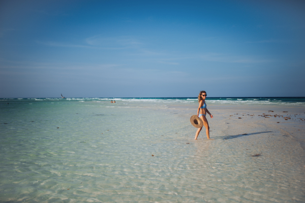 Side view of a beautiful slim woman walking on the beach in a bikini and a straw hat. Full body shot of woman in blue swimsuit, sunglasses enjoying sandy beach and crystalline sea of Mnemba beach in Zanzibar, with copy space. Concept of beach summer vacation.