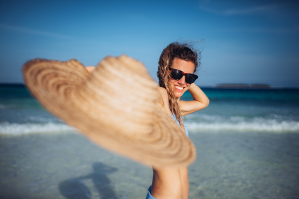 Portrait of a beautiful smiling woman standing on the beach in a bikini with straw hat in hand. Waist up shot of woman in blue swimsuit, sunglasses enjoying sandy beach and crystalline sea of Mnemba beach in Zanzibar. Concept of beach summer vacation.