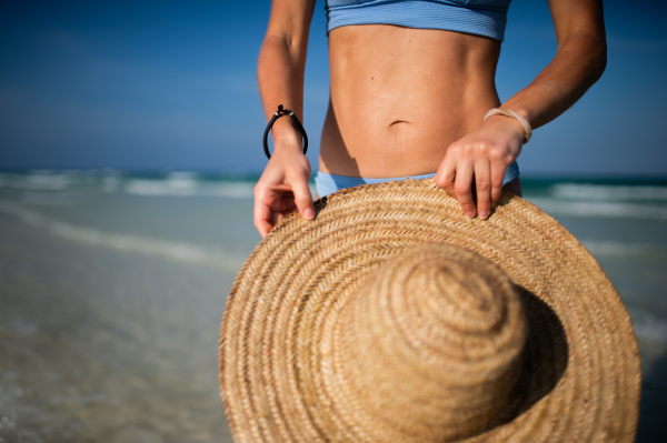 Close up of beautifuly shaped women's midsection, belly, stomach on the beach near the sea. Detail shot of woman in blue bikini on beach with straw hat in hand with copy space. Sandy beach and crystalline sea of Mnemba beach in Zanzibar. Early pregnancy on vacation. Concept of beach summer vacation.