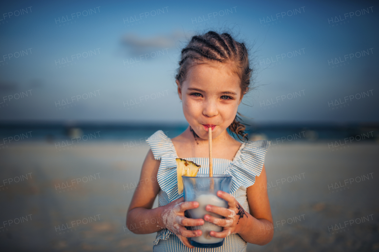 Portrait of a little adorable girl on a sandy beach by the sea. A girl with braids is drinking a non-alcoholic mixed drink. Concept of beach summer vacation with kids.